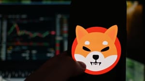 Het logo van de memecoin shiba inu (SHIB) op een mobiele telefoon met op de achtergrond een koers in candle chart format, op een cryptobeurs Het logo van de memecoin shiba inu (SHIB) op een mobiele telefoon met op de achtergrond een koers in candle chart format, op een cryptobeurs